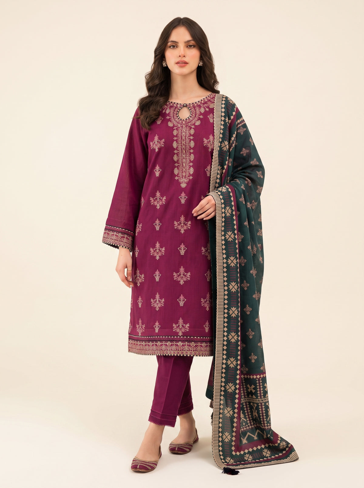 A woman modeling a traditional three-piece salwar kameez suit in deep magenta, featuring intricate gold embroidery on the kurta, matching trousers, and a dark green dupatta with gold and silver patterns.